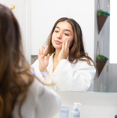 Beautiful young woman in white bathrobe staring at a mirror applying Thalgo skin care cream product to her face.