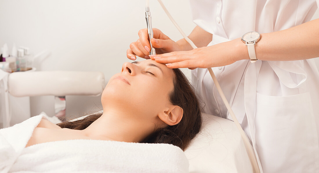 Young woman lying down wearing white robe receiving micro-dermabrasion treatment by esthetician.