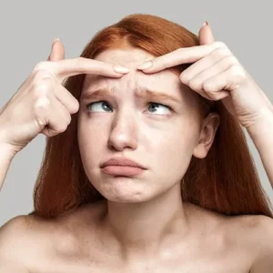 Stop acne worried young redhead woman examining her face while standing against grey background